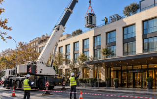 Camión grúa con pluma extendida izando un rack técnico metálico sobre la cubierta de un hotel urbano del Eixample de Barcelona mientras un operario con casco recibe el equipo y otro en la acera dirige la maniobra con señales manuales